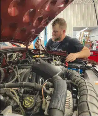 A mechanic works on the engine of a car with the hood raised. He is focused on using a diagnostic tool. Various tools and equipment are visible in the background inside the garage. The engine and its components are prominently visible in the foreground.