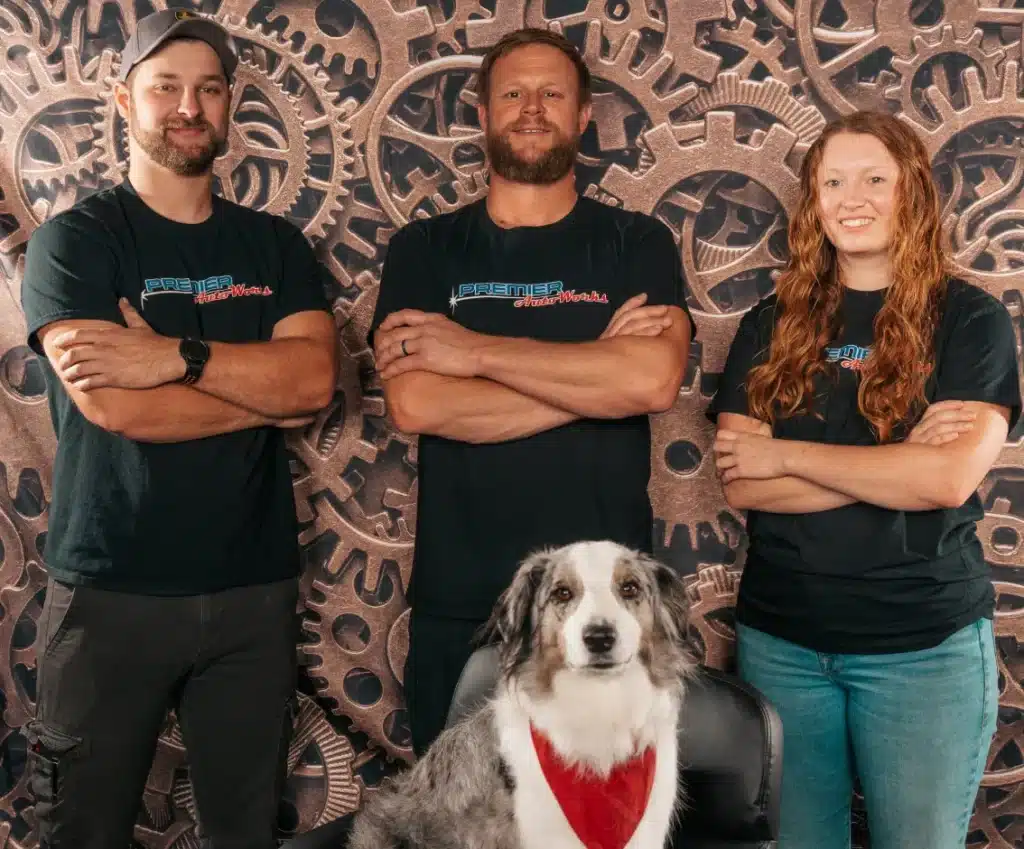 Team members of Premier Auto Works in New Oxford, PA are standing in front of a backdrop with intricate gears. They are all wearing black T-shirts with "Premier Overland" written on them.