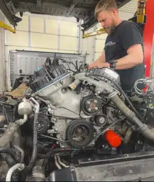 A mechanic is working on a car engine in an auto repair shop. The hood of the car is open, and various tools and parts are visible around him. He is focused on repairing or inspecting the engine components. The background shows a workshop with a garage door.