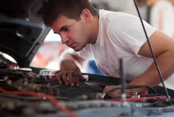 A man in a white t-shirt is focused on examining the engine of a car with the hood open. He seems to be deeply engaged in checking or repairing something under the hood.