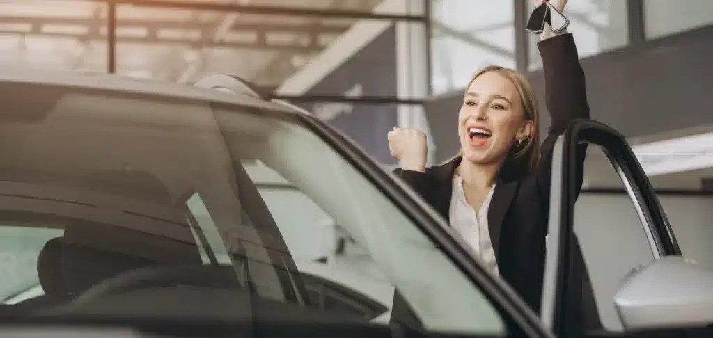 Auto Repair in New Oxford, PA with Premier Auto Works Inc. A woman standing next to a car door is smiling broadly and raising her arm in celebration while holding car keys. She is wearing a blazer, indicating a sense of accomplishment or joy, possibly for buying or leasing a car.