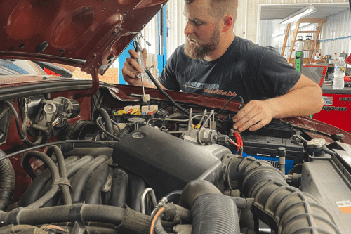 PA State Inspection station in New Oxford, PA. A mechanic works on a car engine in a garage, concentrating on using a tool to adjust a component under the hood.