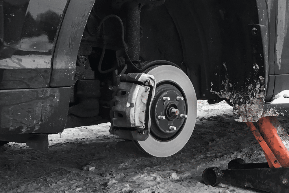 Winter Brake Inspections, Auto repair in New Oxford, PA at Premier Auto Works Inc.. Vehicle lifted on jacks with a missing wheel, exposing the brake rotor and caliper, in a snowy outdoor setting.