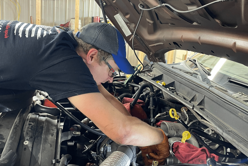 Reliable Mechanic, Auto repair in New Oxford, PA at Premier Auto Works Inc. Mechanic wearing a cap and gloves leans over an open car hood, repairing or inspecting the engine components.