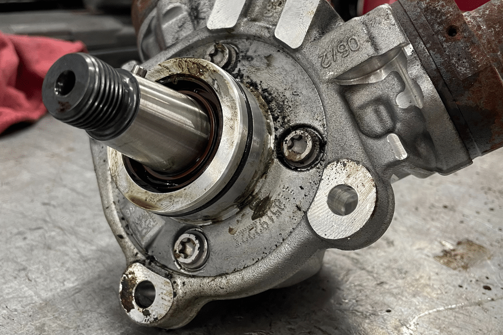 Truck Maintenance Auto repair in New Oxford, PA at Premier Auto Works Inc. A close-up of a metal automotive component, likely a pump or bearing assembly, resting on a workbench. The central shaft protrudes outward, surrounded by a circular seal with visible oil residue and grime. Several mounting bolt holes and fasteners are built into the housing, which shows signs of wear and light corrosion.